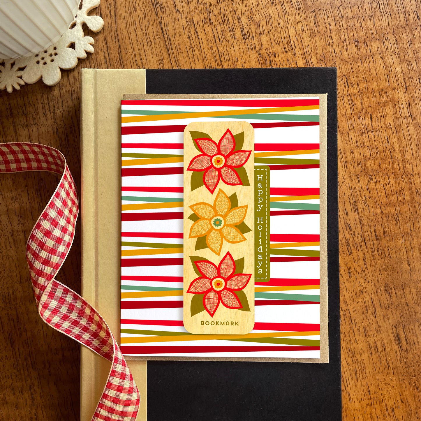 Folded holiday greeting card with colorful red, green and gold stripes, “Happy Holidays” text and a removable birch wood bookmark featuring three red and gold poinsettia flowers. Pictured with a kraft envelope and sitting atop a book on a wooden table with a red gingham ribbon.
