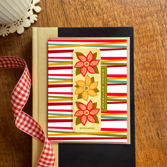 Folded holiday greeting card with colorful red, green and gold stripes, “Happy Holidays” text and a removable birch wood bookmark featuring three red and gold poinsettia flowers. Pictured with a kraft envelope and sitting atop a book on a wooden table with a red gingham ribbon.