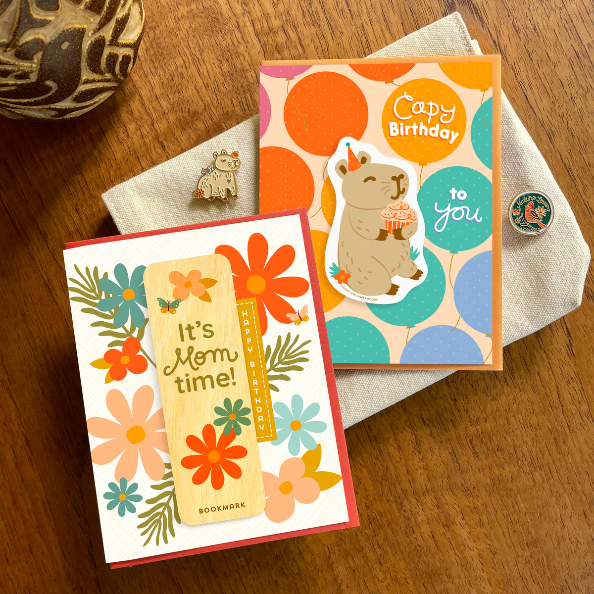 Two handmade greeting cards by Night Owl Paper Goods. One card says "It's Mom time!" with floral illustrations and a removable real wood bookmark with "Happy Birthday"; the other has a colorful polka-dot background with a removable vinyl sticker of an adorable tan capybara sitting and wearing a party hat holding a cupcake and the words "Capy Birthday to you."