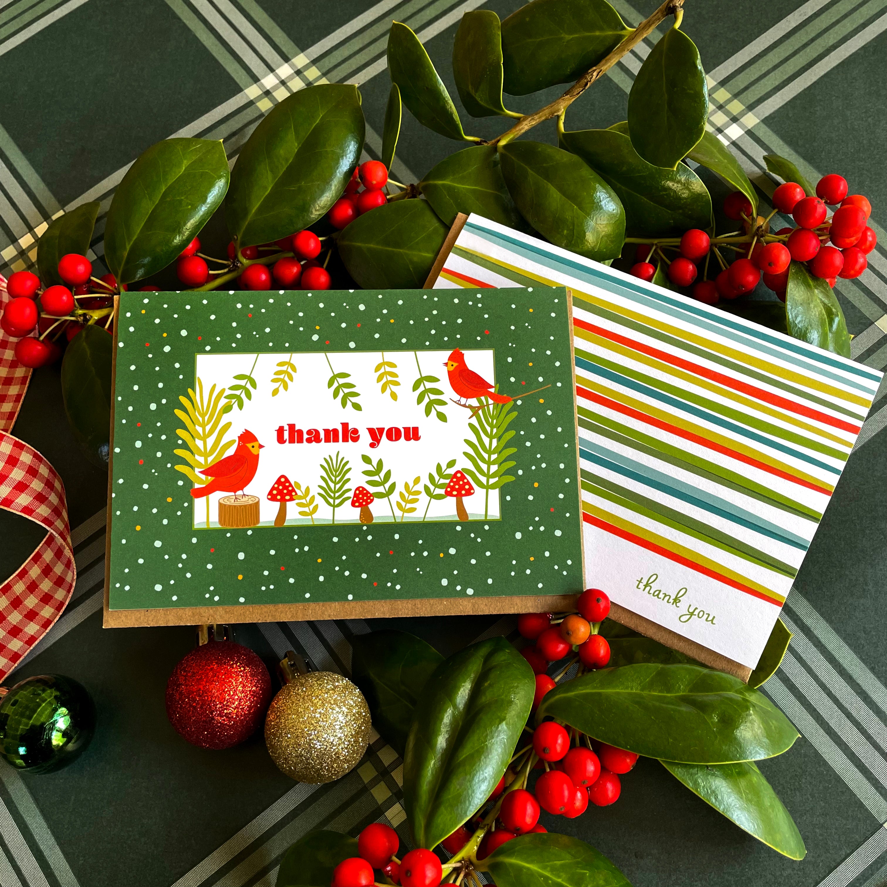Two colorful thank you cards with 'thank you' text surrounded by holly leaves and berries on a plaid background.