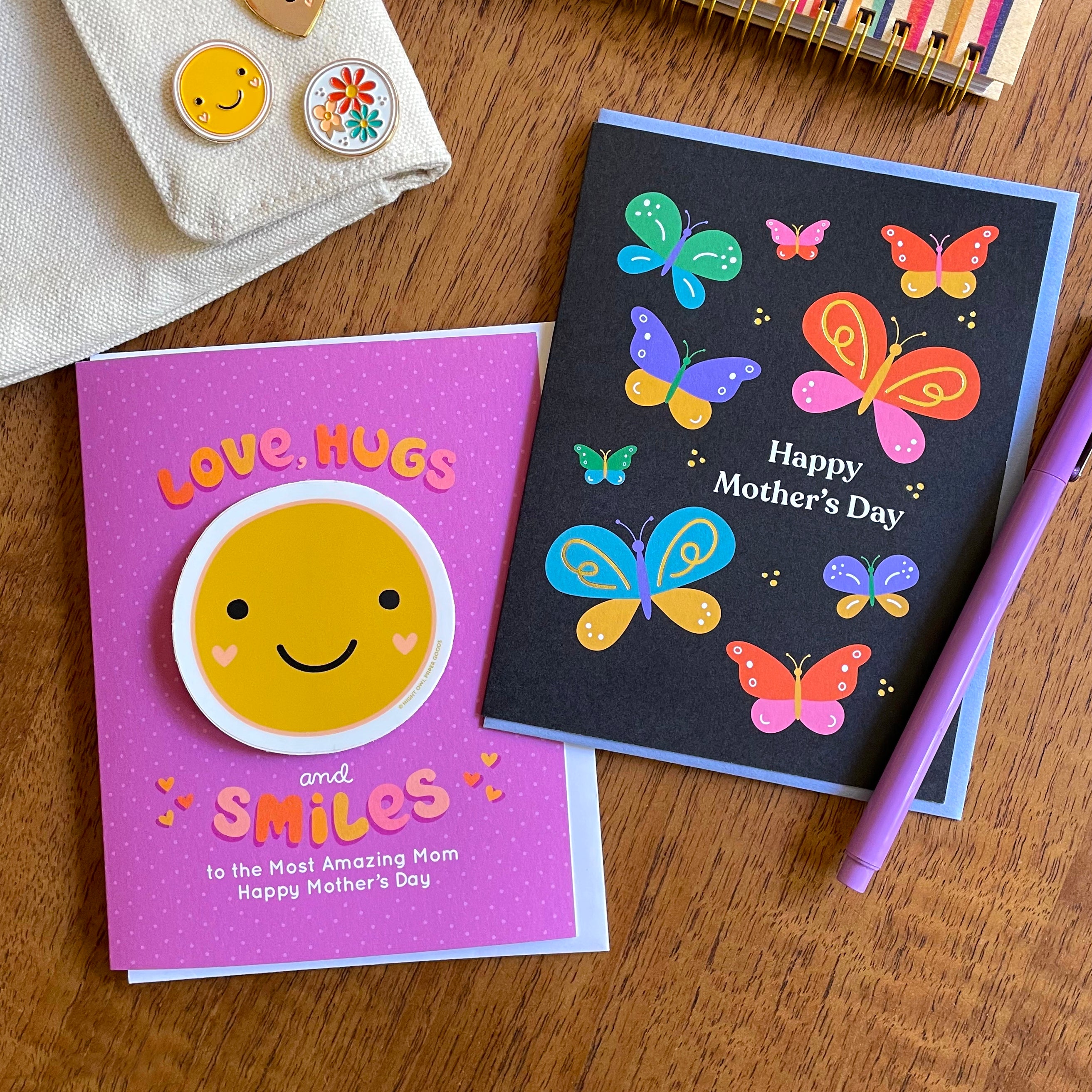 Two Mother's Day greeting cards on a wooden surface with a pen and decorative items.