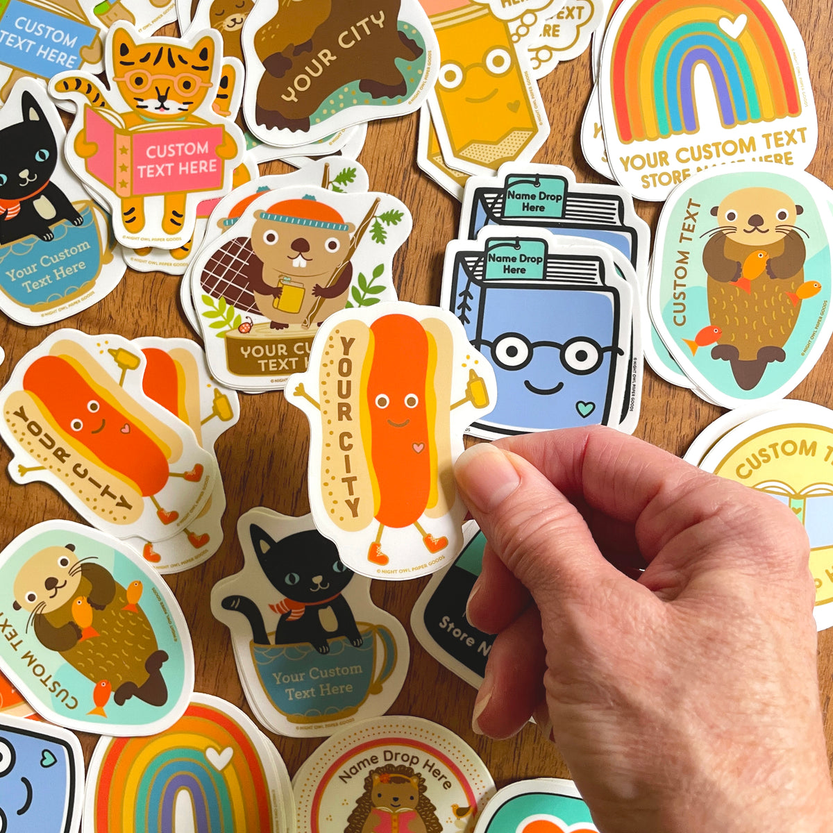 Night Owl Paper Goods custom vinyl stickers. A hand holds a die-cut sticker featuring a smiling hot dog with the text 'YOUR CITY' to show customization option. More stickers feature happy book with eye glasses, otter holding a fish with custom text, black cat in a cup, rainbow with heart, reading hedgehog, beaver on a stump holding a book.