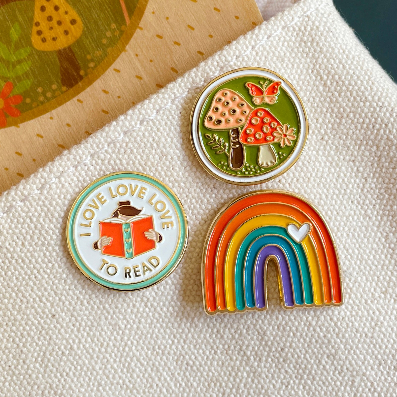 Three colorful pins featuring mushroom design, 'I love to read' design, and rainbow design on a canvas bag.