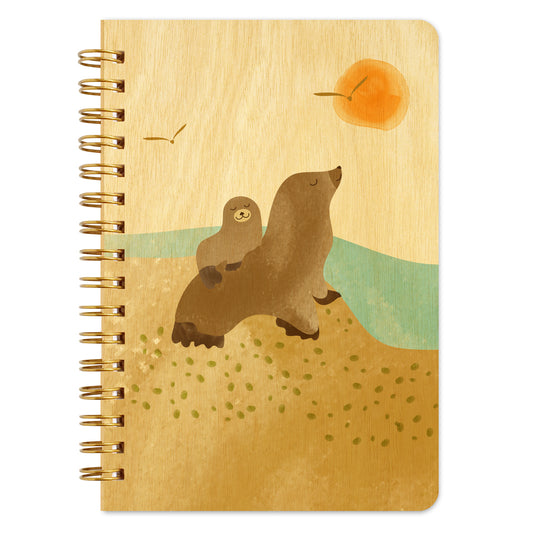 Spiral-bound wood notebook with birch wood covers featuring a sea lion pup riding on its mother’s back beside the ocean with seagulls and a setting sun.