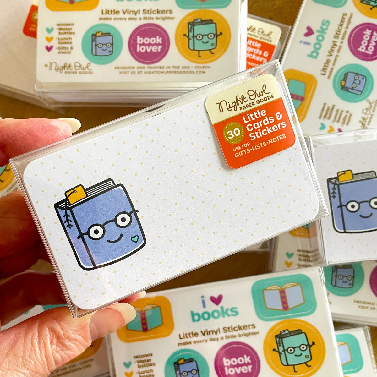 A hand holds a clear box of Night Owl Little Cards & Stickers with a smiling blue book design on a white card. The label on the box reads, "30 - Little Cards & Stickers - Use for Gifts - Lists - Notes." Background shows an array of boxes of the same product.