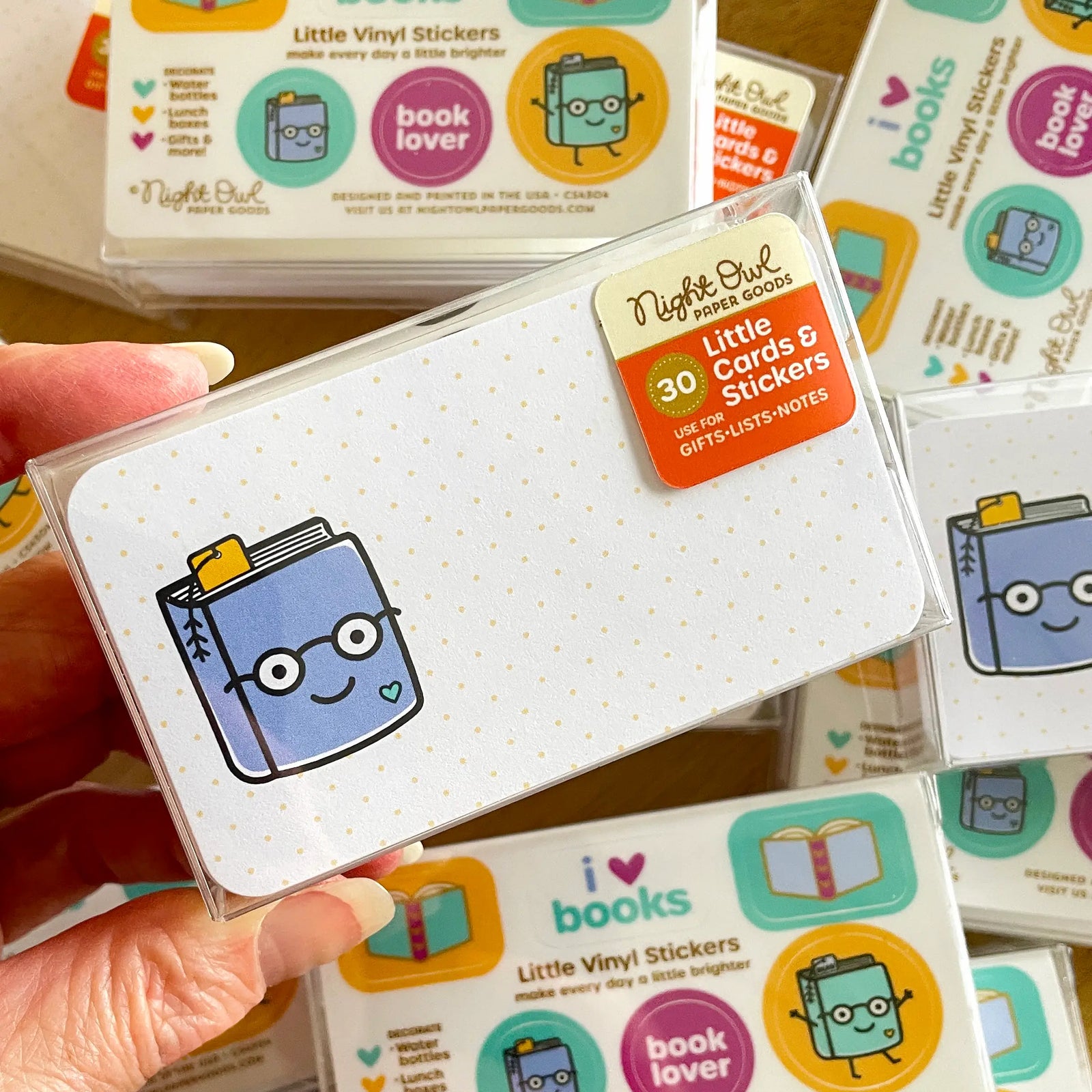 A hand holds a clear box of Night Owl Little Cards & Stickers with a smiling blue book design on a white card. The label on the box reads, "30 - Little Cards & Stickers - Use for Gifts - Lists - Notes." Background shows an array of boxes of the same product.