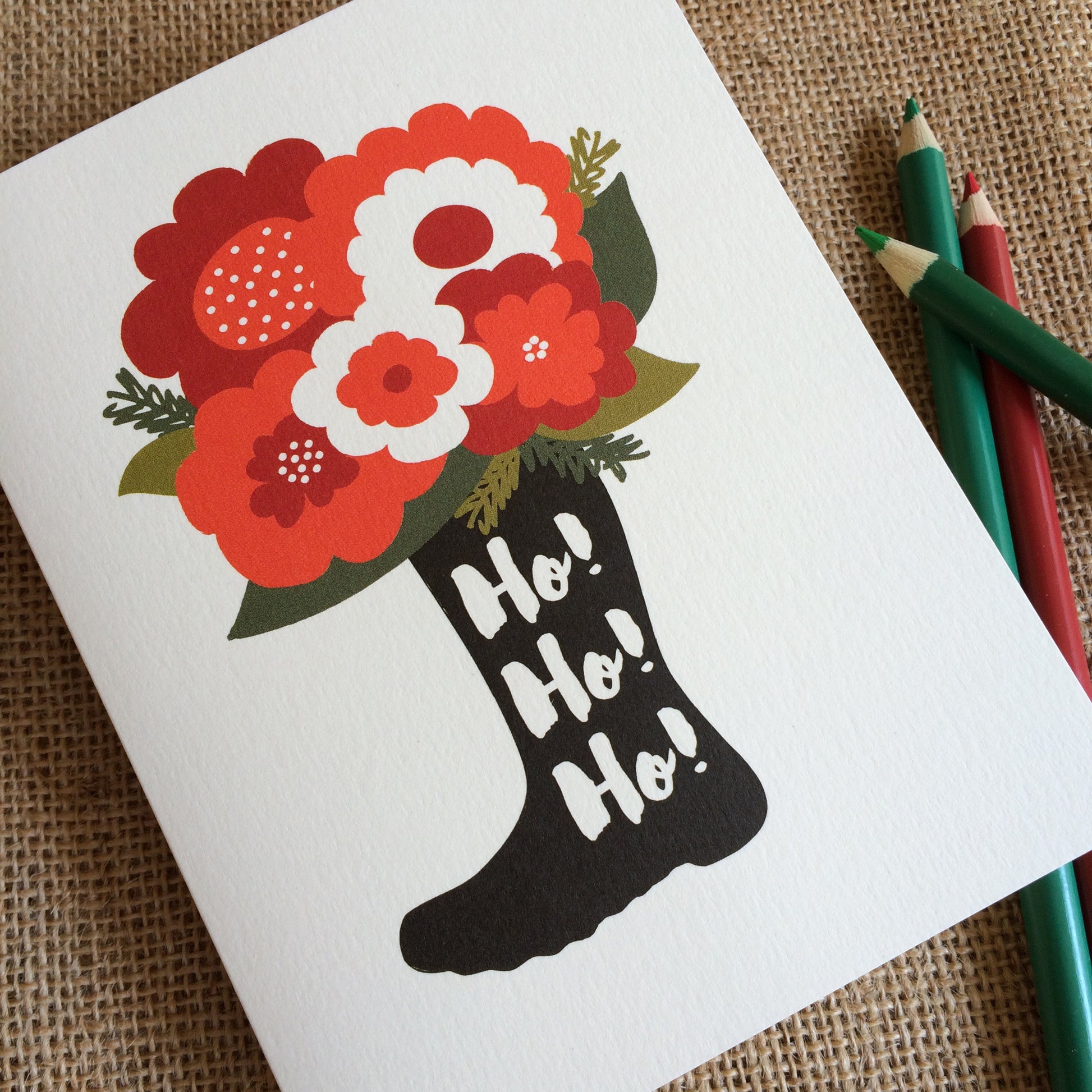 Front of a folded holiday card with flowers blooming out the top of a black boot with Ho! Ho! Ho! text on an off-white background.