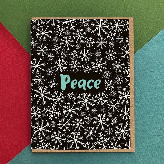 Black holiday greeting card with a playfully illustrated snowflake pattern and 'Peace' text. Background of photo is colorful.