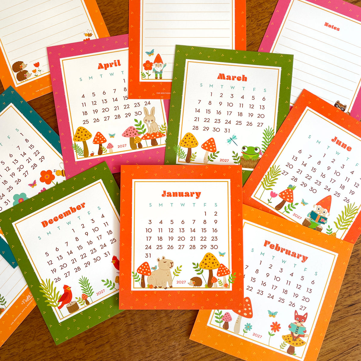 2027 Forest Friends desk calendar with multiple pages showing Night Owl Paper Goods woodland animal designs. Critters include a capybara with blue bird on its back, happy gnome reading, green frog with tongue catching a dragonfly, bunny rabbit sitting on a red mushroom, reading fox sitting on a mushroom.
