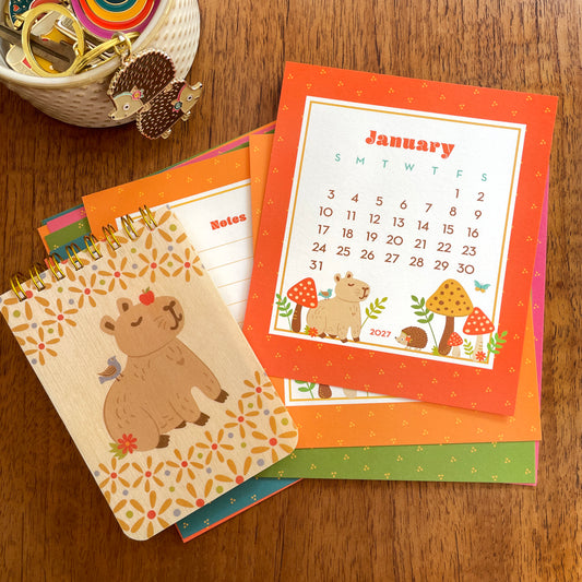 2027 Forest Friends desk calendar pages featuring cute Night Owl Paper Goods woodland animals as well as a mini notepad with wood covers with an adorable capybara sitting with a blue bird on its back and an apple on its nose. Also featured is a gold enamel keychain with two colorful hedgehogs wearing flower headbands.
