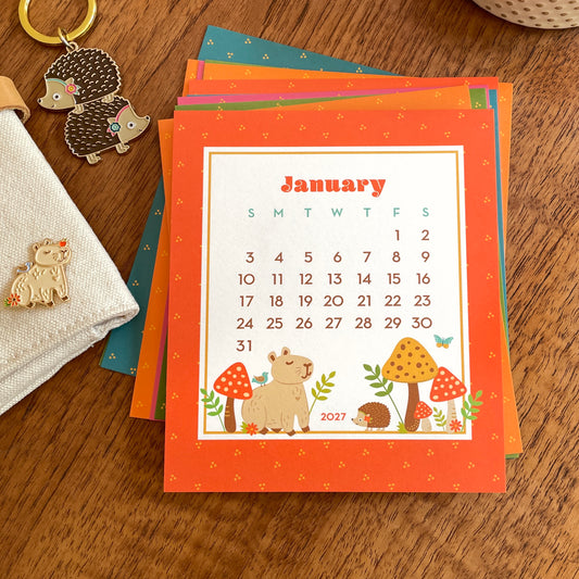 2027 Forest Friends desk calendar with January page featuring signature Night Owl Paper Goods woodland animals. Critters include a capybara with blue bird on its back and colorful mushrooms and a cute hedgehog. Coordinating accessories include gold trim enamel capybara enamel pin and two hedgehogs in a stack keychain.