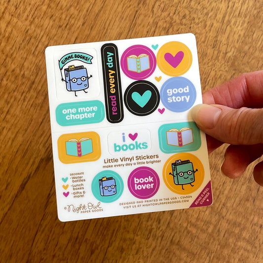 Hand holding a sheet of colorful book-themed kiss-cut vinyl stickers featuring cute book characters, open books, hearts and sayings like “Gimme Books!”, “One More Chapter,” “Read Every Day,” “Good Story” and “Book Lover” in aqua, purple, yellow, black and blue. Text on the sheet reads “Little Vinyl Stickers - make every day a little brighter - Decorate water bottles, lunch boxes, gifts & more - Night Owl Paper Goods - Designed and printed in the USA.”