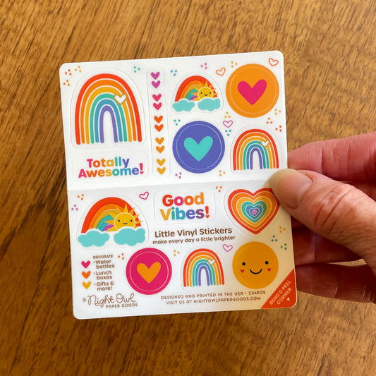 Hand holding a sheet of colorful rainbow-themed kiss-cut vinyl stickers on a white background. The stickers feature rainbows, hearts, a smiling sun, a rainbow heart, a happy face and sayings like "Totally Awesome!" and "Good Vibes!" in bright colors like orange, yellow, teal, blue and pink. Text on the sheet reads "Little Vinyl Stickers - make every day a little brighter - Decorate water bottles, lunch boxes, gifts & more - Night Owl Paper Goods - Designed and printed in the USA"