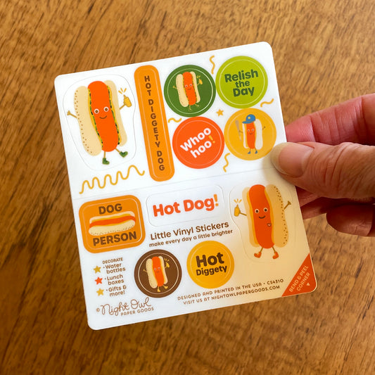 Hand holding a sheet of colorful kiss-cut vinyl stickers on a white background featuring a cheerful hot dog, hot dog illustrations, a baseball-themed hot dog and sayings including "Dog Person" "Hot Diggety Dog," "Relish the Day," "Whoo hoo," "Hot Dog!" and "Hot Diggety." The stickers use bright orange, yellow, green, brown and red colors. Text on the sheet reads "Little Vinyl Stickers - make every day a little brighter - Decorate water bottles, lunch boxes, gifts & more - Night Owl Paper Goods - Designed an