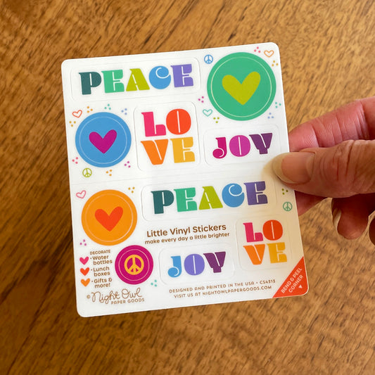 Hand holding a sheet of colorful kiss-cut vinyl stickers on a white background. The stickers feature hearts, peace signs and “PEACE LOVE JOY” designs in various shades of vibrant teal, green, blue, orange, pink, purple and yellow. Text on the sheet reads “Little Vinyl Stickers - make every day a little brighter - Decorate water bottles, lunch boxes, gifts & more - Night Owl Paper Goods - Designed and printed in the USA.”