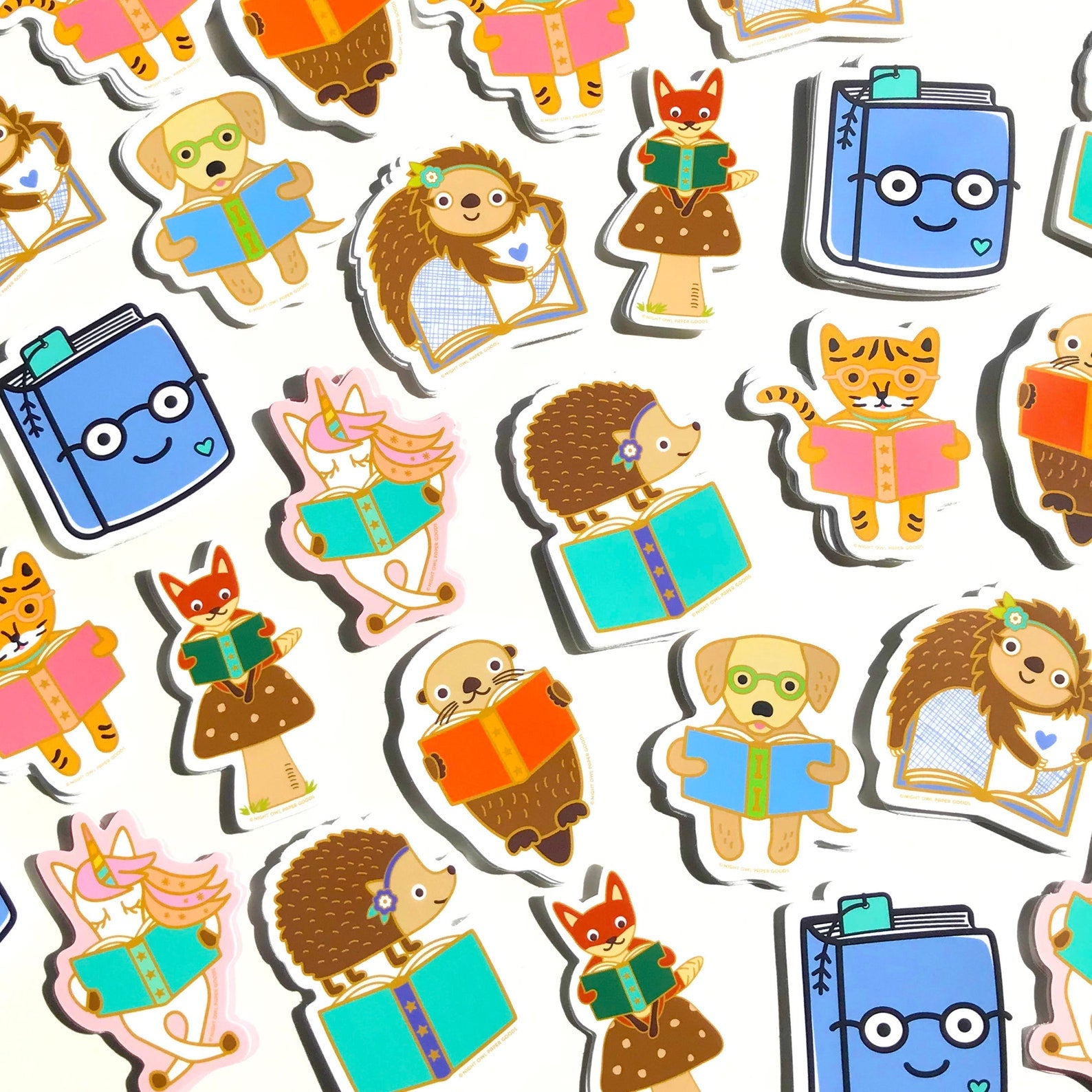Array of book lover stickers, including a reading golden retriever, a reading sloth, a reading unicorn, a reading otter, a hedgehog standing on a book, an otter holding a book, a reading tiger and a happy book wearing glasses.