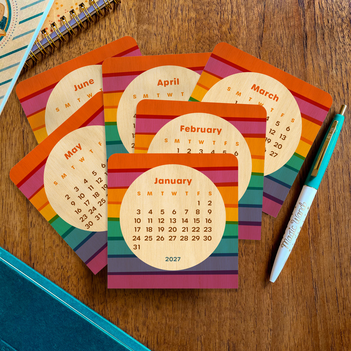 Birch wood mini desk calendar pages arranged on a wooden desk, showing colorful rainbow stripes and several 2027 months, including January, February, March, April, May and June.