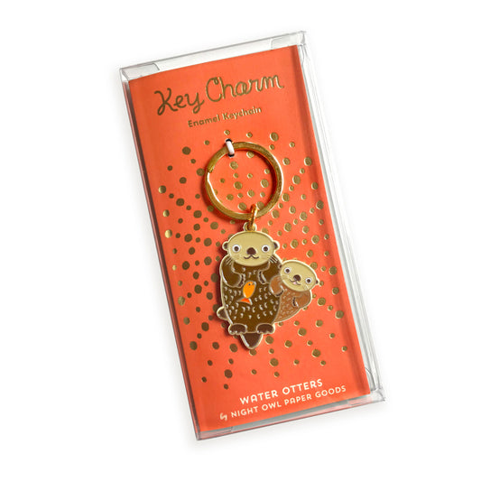 Otter enamel keychain on gold foil stamped orange backer card packaged in a clear gift box. Backer card reads "Key Charm Enamel Keychain" at top and "Water Otters by Night Owl Paper Goods" at bottom.