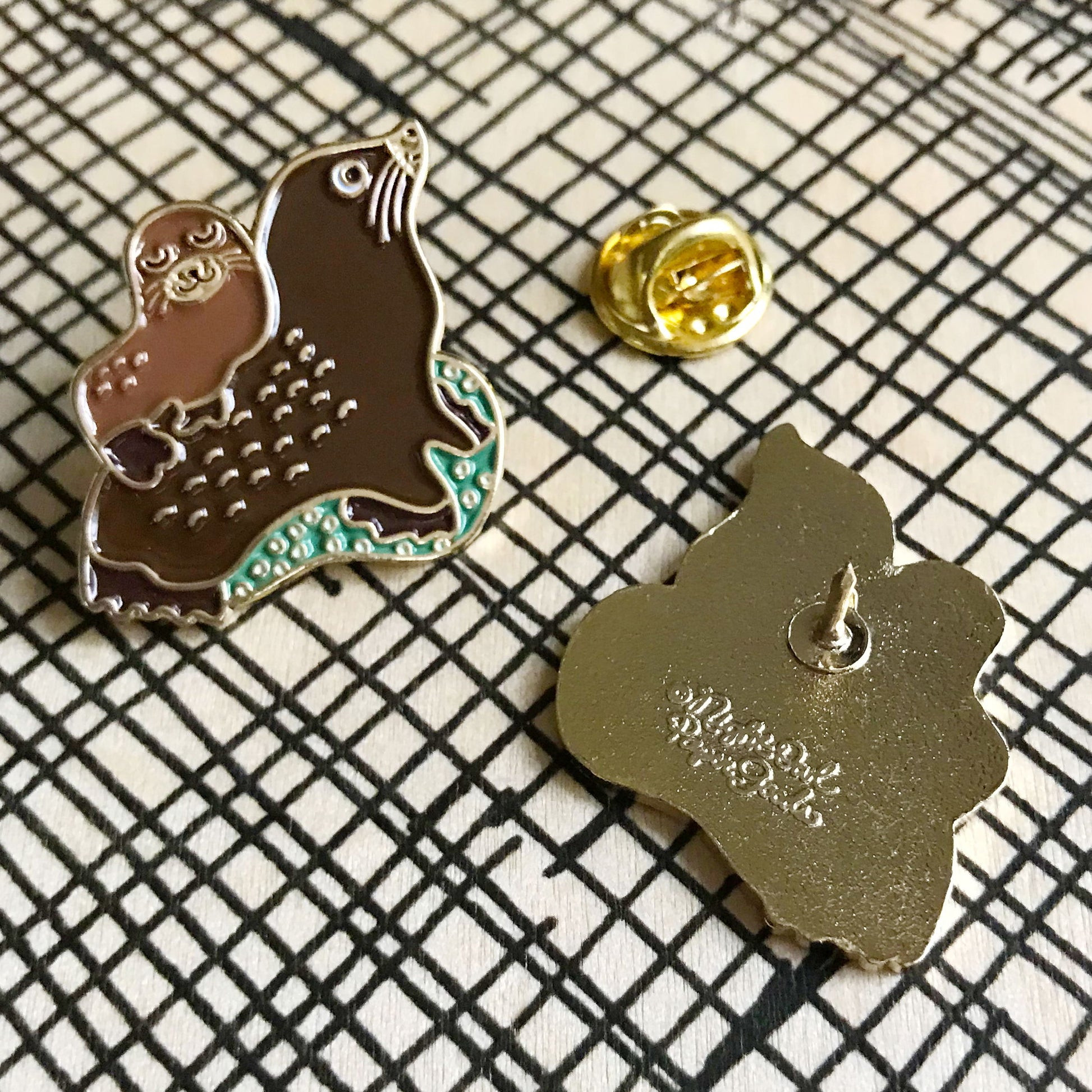 Front and back of an enamel pin of a sea lion with a smaller sea lion on its back. Back shows Night Owl Paper Goods script logo.