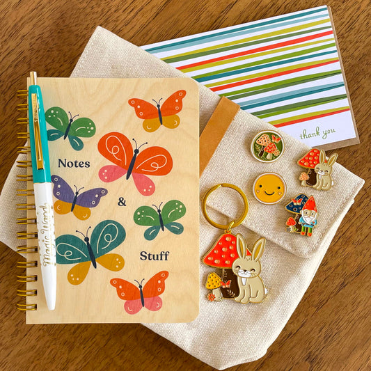Butterfly wood notebook, Magic Wand pen, Lively Stripes Thank You card and Rabbit keychain alongside Mushroom, Rabbit, Smiley Face and Gnome enamel pins. 