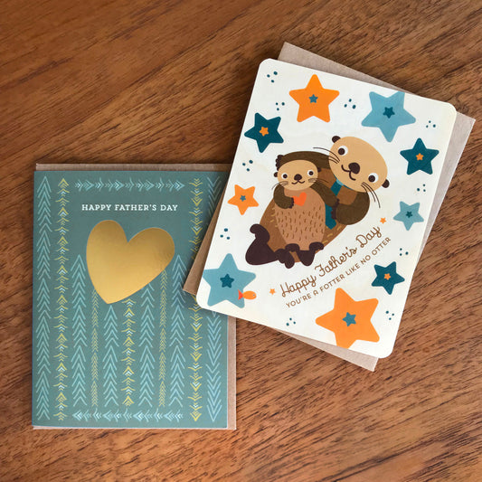 Gold Heart Dad Foil Stamped Father's Day card and Otter Dad Wood Father's Day card, both pictured with kraft envelopes on a wood table.