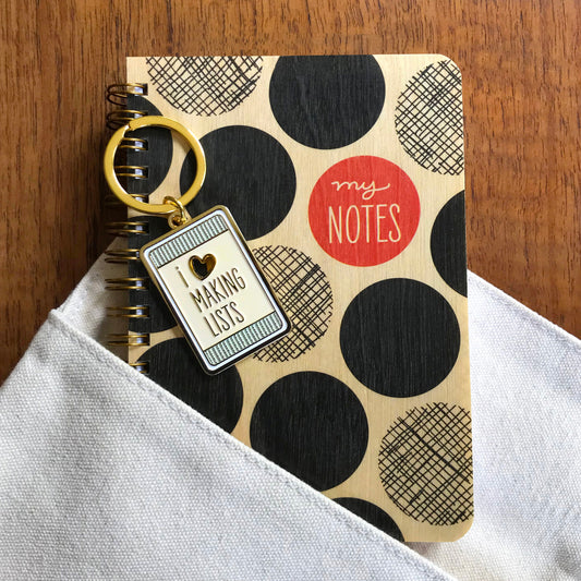 Making Lists keychain sitting on top of a Big Dots Notes wood notebook emerging from a canvas pocket.