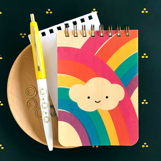 Spiral-bound wood mini notepad with birch wood covers featuring a smiling cloud nestled between two bold crossing rainbows in vivid red, orange, yellow, green and purple tones. Next to it sits a ballpoint click pen and a torn out sheet of blank white inner paper. Click pen features fun illustrations of happy sun and shooting stars. 