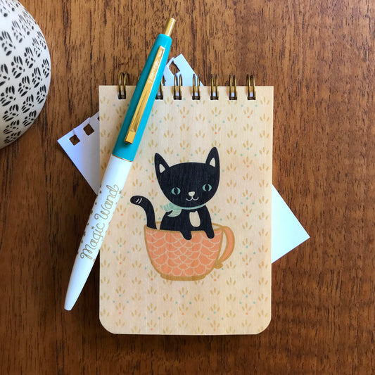 Spiral-bound wood mini notepad with birch wood covers featuring a black cat with green eyes sitting inside a pink teacup alongside an aqua and white Magic Wand ballpoint pen.