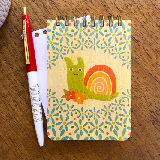 Spiral-bound wood mini notepad with birch wood covers featuring a smiling snail surrounded by a bold retro floral pattern sits upon a blank white inner page that has been torn out and alongside a red and white Getting Stuff Done ballpoint pen.