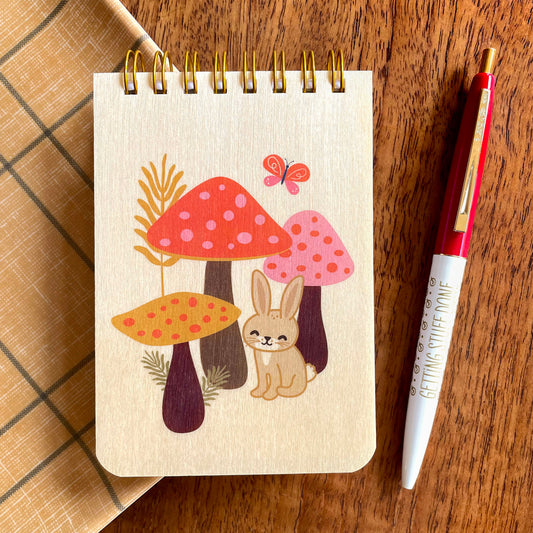Spiral-bound wood mini notepad with birch wood covers featuring a happy bunny nestled among spotted mushrooms with a butterfly and wildflowers alongside a Getting Stuff Done ballpoint pen.