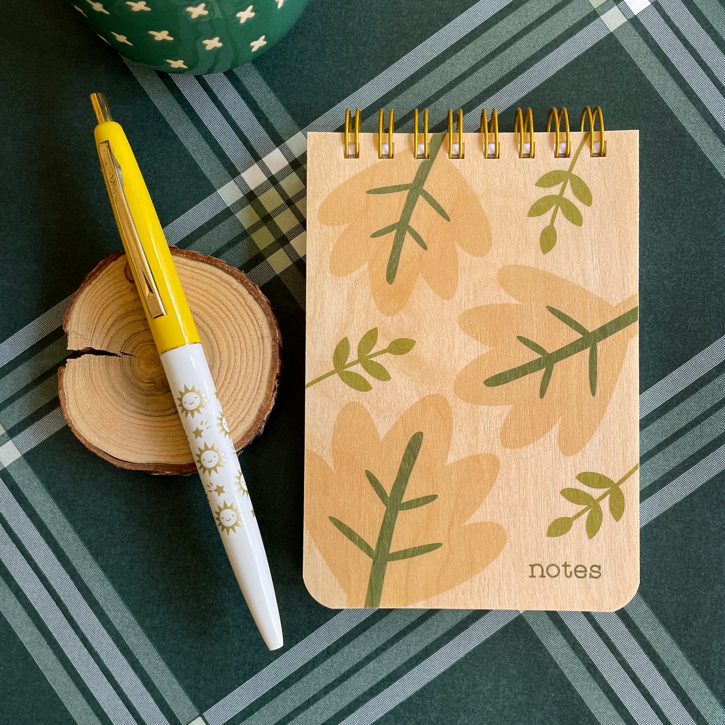 Mini wood notepad with big neutral color leave and green stems. Notebook says 'notes' on bottom right corner. Click pen with yellow cap and white barrel with gold stars and happy suns.