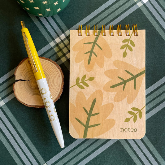 Mini wood notepad with big neutral color leave and green stems. Notebook says 'notes' on bottom right corner. Click pen with yellow cap and white barrel with gold stars and happy suns.