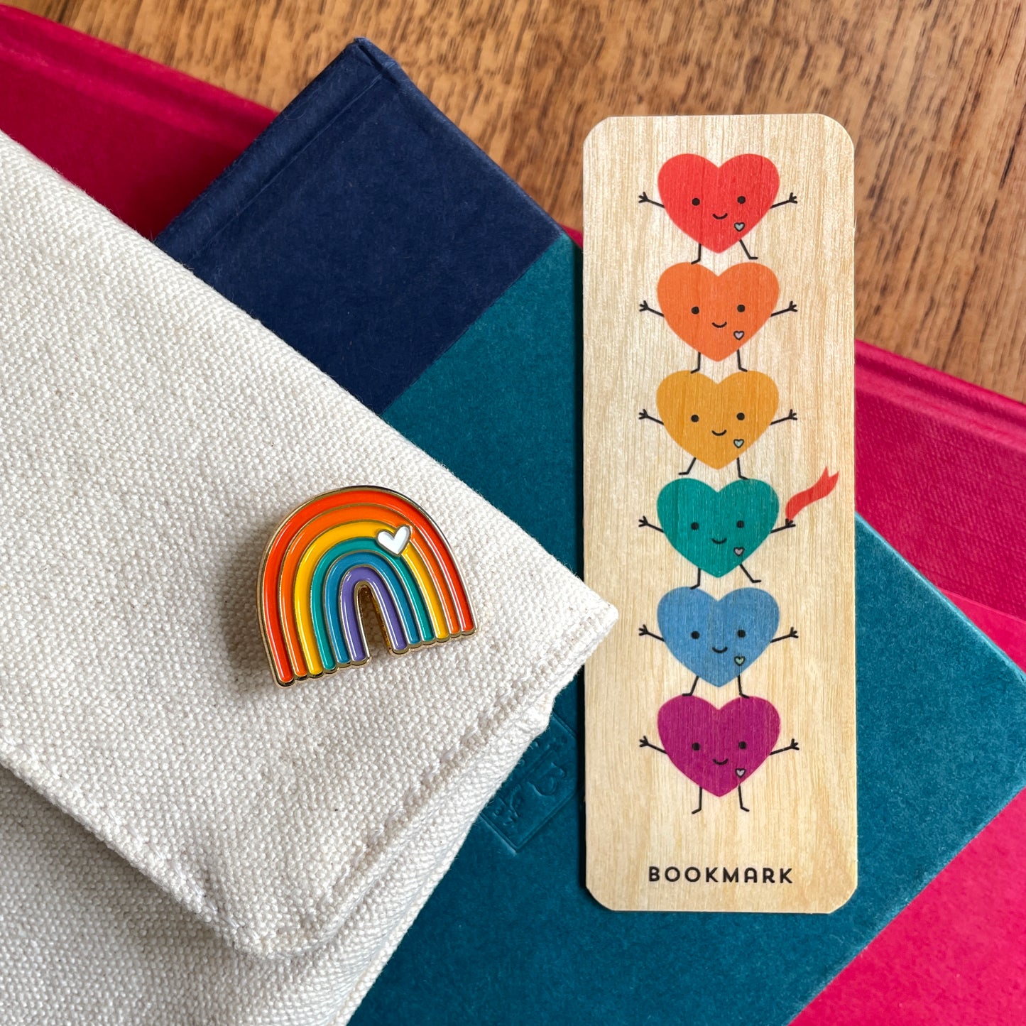A wooden bookmark with six colorful smiling hearts and the word 'BOOKMARK' at the bottom, displayed next to a rainbow enamel pin on a folded white fabric surface with books in the background.