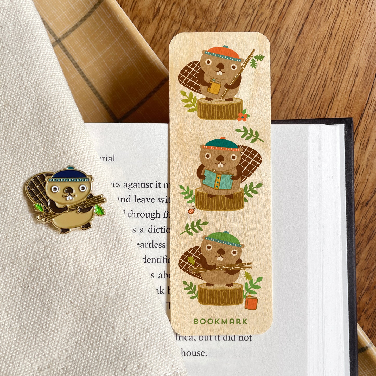 A wooden bookmark featuring three beavers each holding a book, displayed on an open book next to a matching beaver enamel pin on a textured cream fabric.