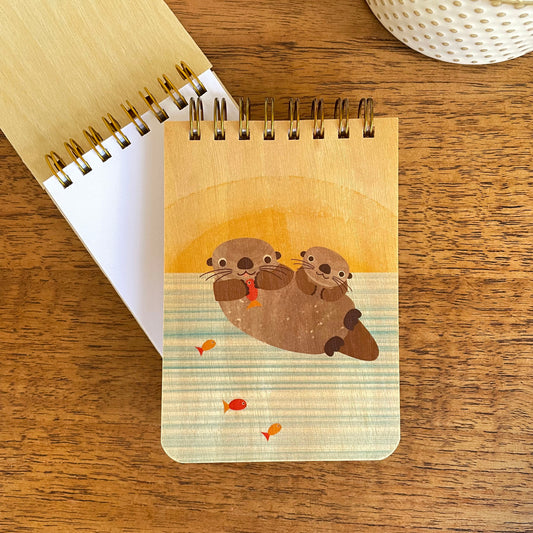 Spiral-bound wood mini notepad with birch wood covers featuring two otters floating together against a warm golden sunset with small orange fish swimming nearby sits upon an open mini notepad that shows blank white inner pages.