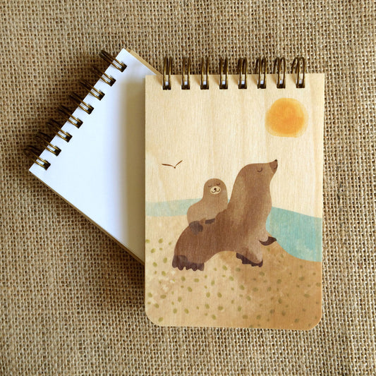 Spiral-bound wood mini notepad with birch wood covers featuring two sea lions of different sizes resting together on a sunny sandy shore with a seagull and golden sun overhead sits upon an open mini notepad that shows blank white inner pages.