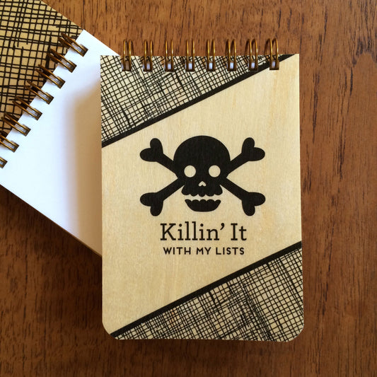 Spiral-bound wood mini notepad with birch wood covers, skull and crossbones design and "Killin' It With My Lists" text sits upon an open mini notepad that shows blank white inner pages.