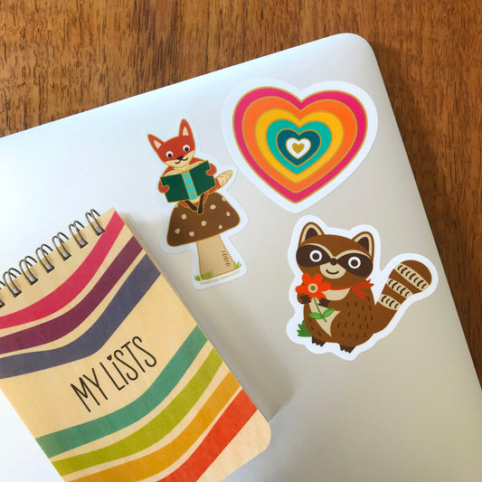 Mini lists notepad and laptop computer with fun stickers of colorful rainbow heart, fox reading on a mushroom and a raccoon holding a flower. 