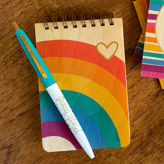 Mini notepad with birch wood covers and spiral binding on top edge. Rainbow design and heart symbol.  Accompanied by a click-top pen with aqua top and gold Magic Wand text.