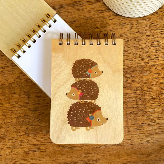 Spiral-bound wood mini notepad with birch wood covers featuring three hedgehogs stacked on top of each other, each wearing a colorful floral accessory sits upon an open mini notepad that shows blank white inner pages.