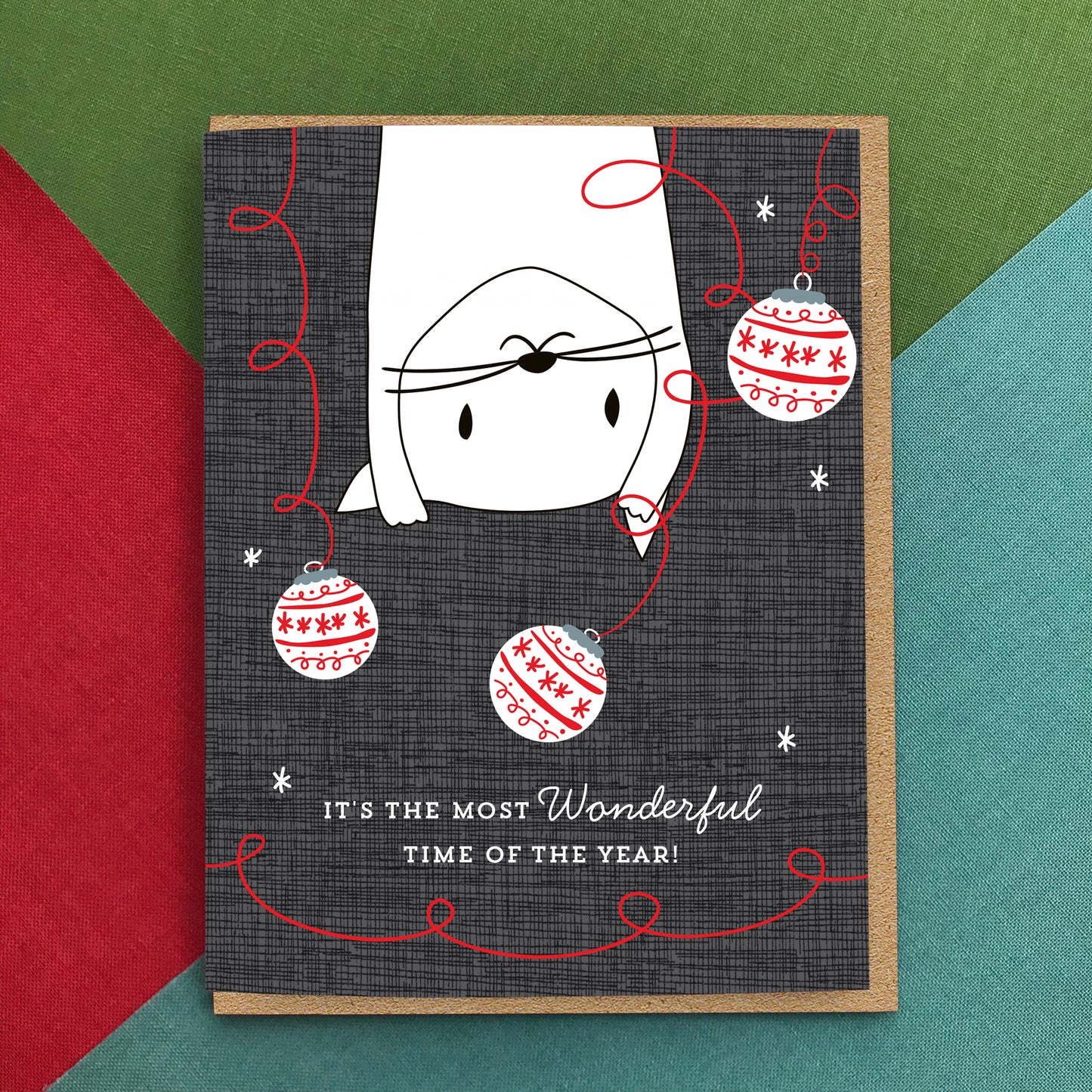 Holiday card pictured on colorful background. Card has a white illustrated cat swinging from the top of the card surrounded by Christmas ornaments on a dark textured background. Text reads 'It's the most wonderful time of the year!'