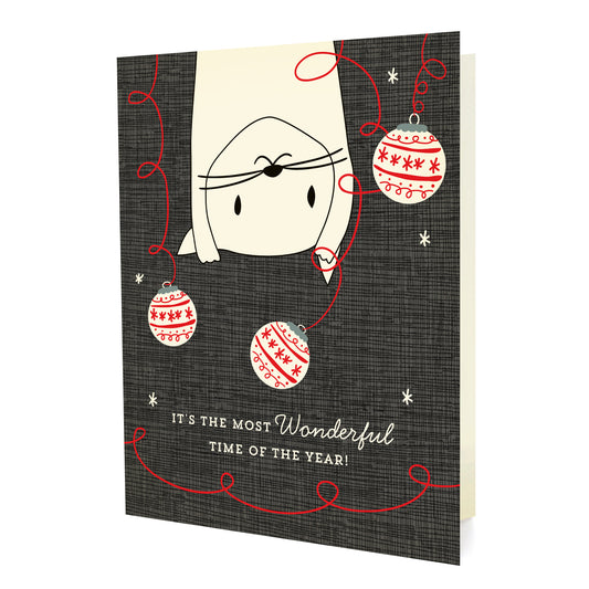 Front of folded holiday card featuring a playful cat swinging from the top among Christmas ornaments with “It’s the most wonderful time of the year!” text on a textured background.