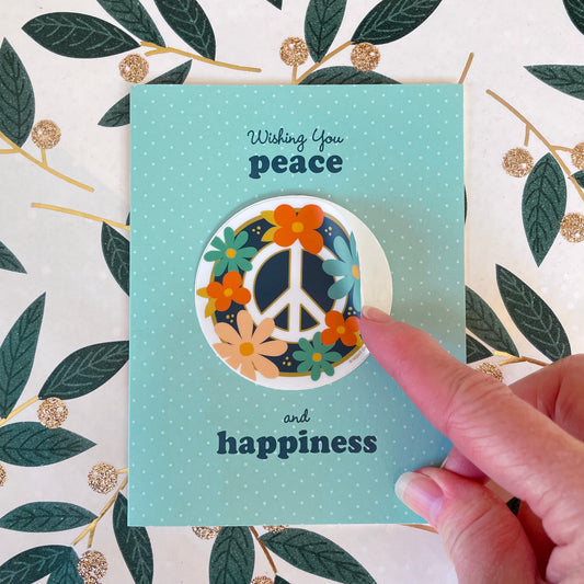 Blue holiday card featuring a removable sticker with peace symbol and flowers. Card sentiment reads "Wishing you peace and happiness". Hand is showing how the sticker can be removed.