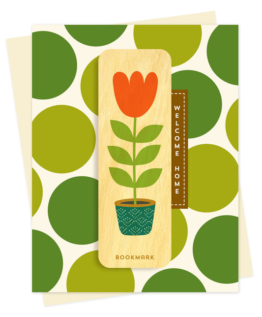 Folded new home card with a cream background and bold green polka dot pattern, with Welcome Home text and a removable birch wood bookmark featuring an orange tulip in a teal patterned pot. Pictured with a cream envelope.