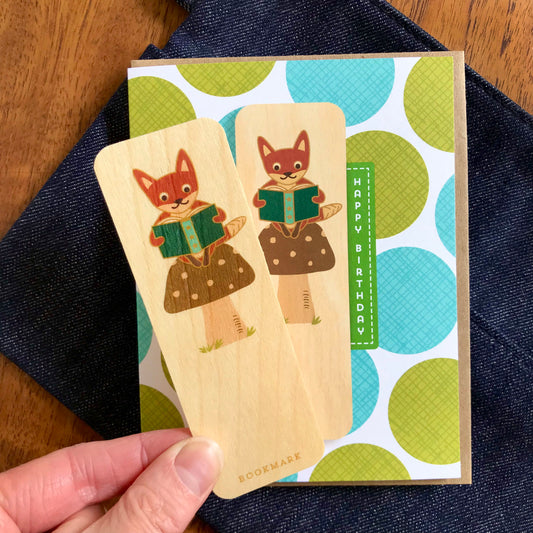 Detail of a fox wood bookmark being removed from a folded birthday greeting card with a white background and oversized green and teal polka dot pattern, Happy Birthday text. The wood bookmark features a fox reading a book while perched on a mushroom. Card is pictured with a kraft envelope.