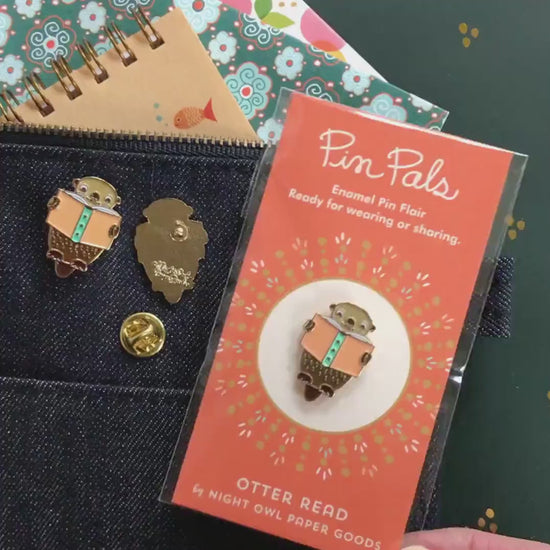 Front of enamel pin is an otter reading a book with gold accents. Back of pin is gold with the Night Owl Paper Goods script logo. Pin is mounted on a backer card that says "Pin Pals - Enamel Pin Flair - Ready for wearing or sharing - OTTER READ - by Night Owl Paper Goods" and is packaged in a clear bag.