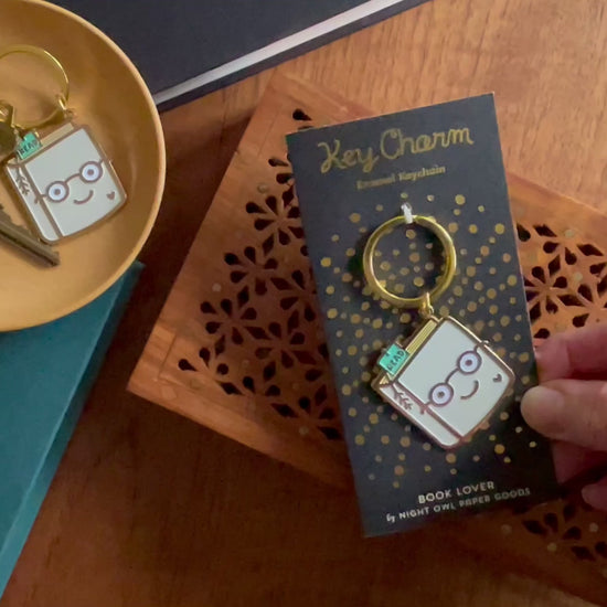 Front of enamel keychain is a smiling book wearing glasses with gold accents. Keychain is mounted on a backer card that says "Key Charm - Enamel Keychain - BOOK LOVER by Night Owl Paper Goods" and is packaged in a clear box.