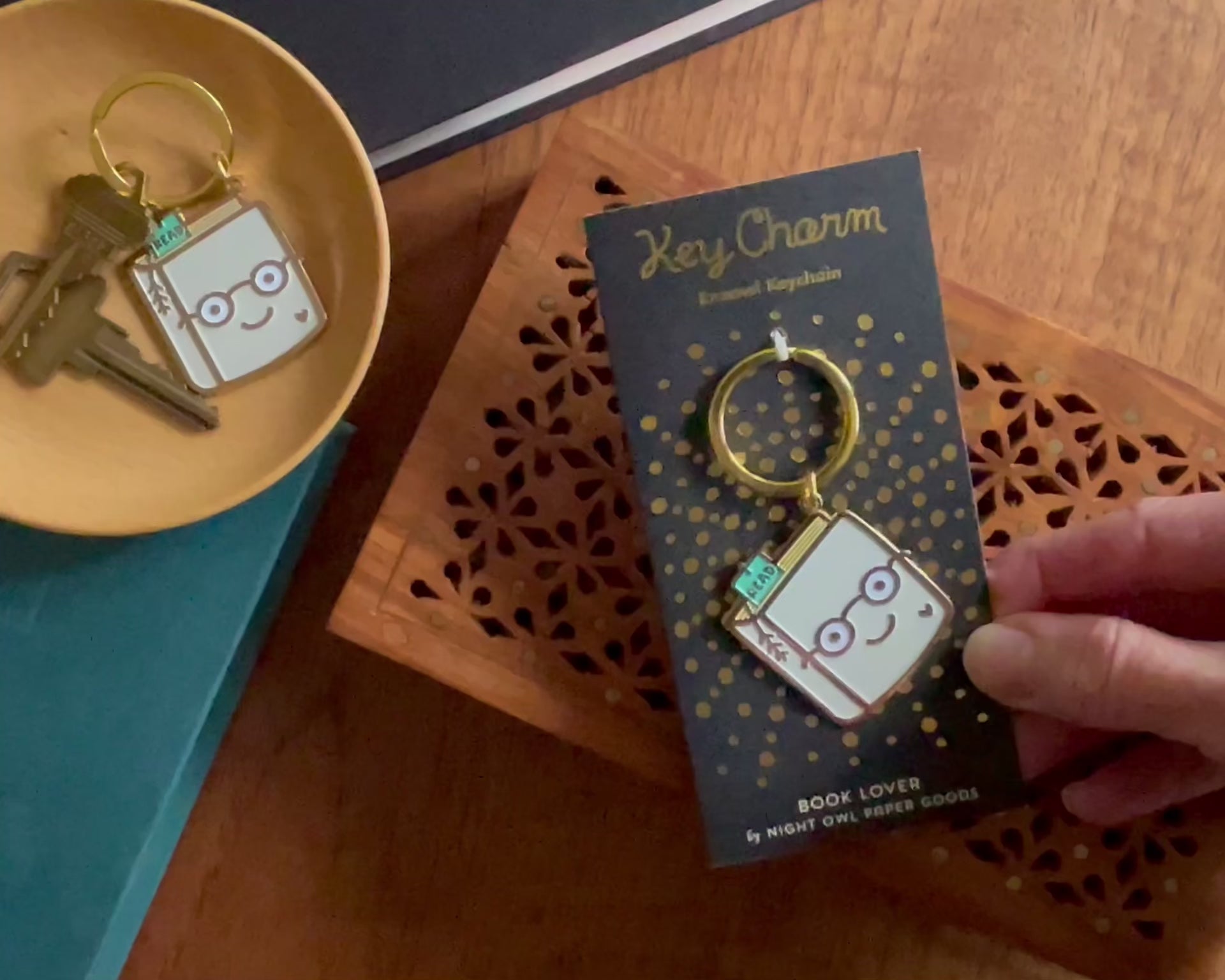 Front of enamel keychain is a smiling book wearing glasses with gold accents. Keychain is mounted on a backer card that says "Key Charm - Enamel Keychain - BOOK LOVER by Night Owl Paper Goods" and is packaged in a clear box.
