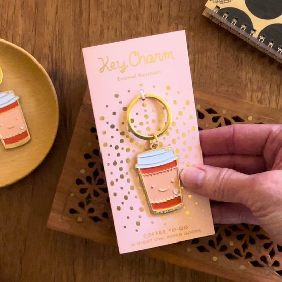 Front of enamel keychain is a smiling coffee cup with gold accents. Keychain is mounted on a backer card that says "Key Charm - Enamel Keychain - COFFEE TO GO by Night Owl Paper Goods" and is packaged in a clear box.