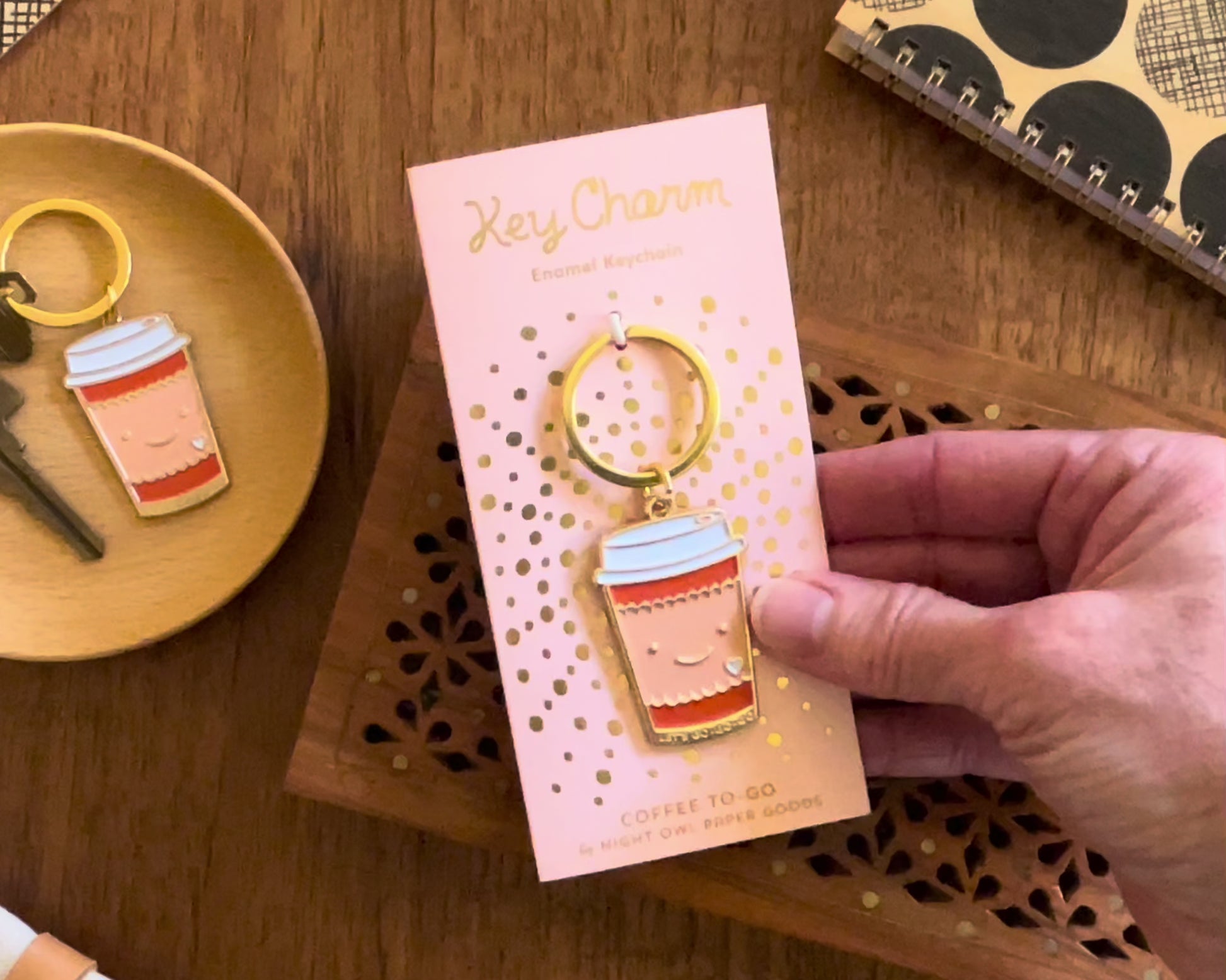 Front of enamel keychain is a smiling coffee cup with gold accents. Keychain is mounted on a backer card that says "Key Charm - Enamel Keychain - COFFEE TO GO by Night Owl Paper Goods" and is packaged in a clear box.